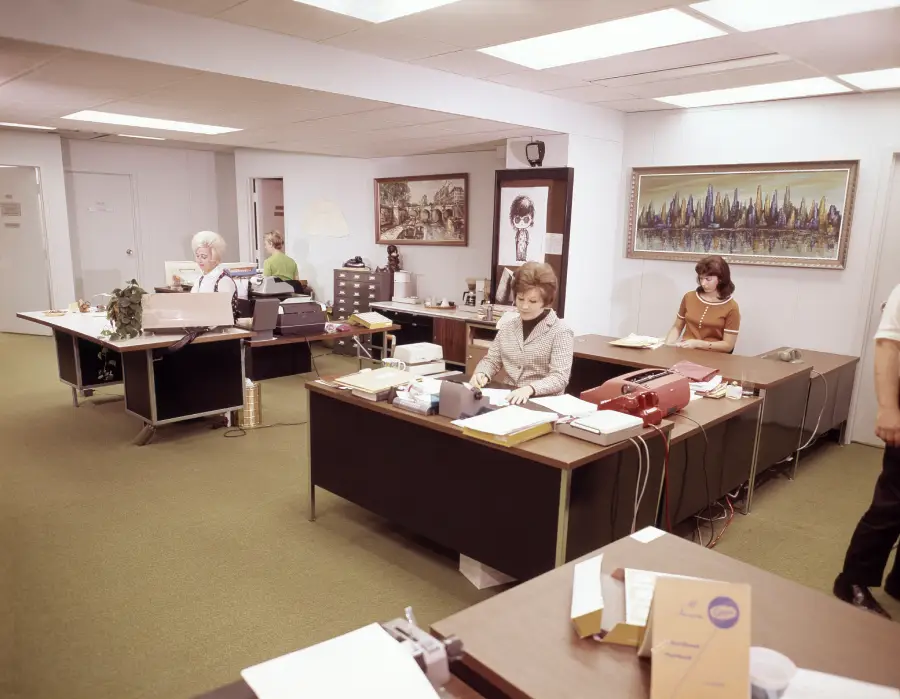 Four women work at desks with typewriters in a vintage office. The room has art on the walls, green carpet, and office supplies. A partial view of a person standing is on the right. The scene suggests a typical 1970s workplace.