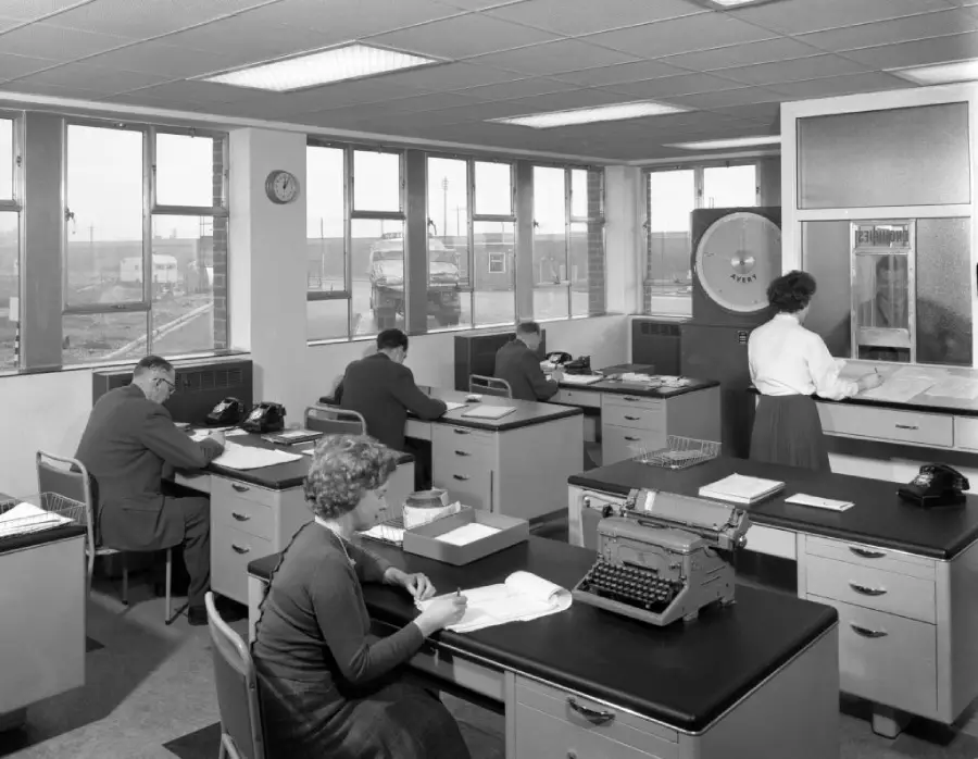 Black-and-white photo of a 1950s office with men and women working at desks. There are typewriters, papers, telephones, and a large computer in the background. The room has large windows and fluorescent lights.
