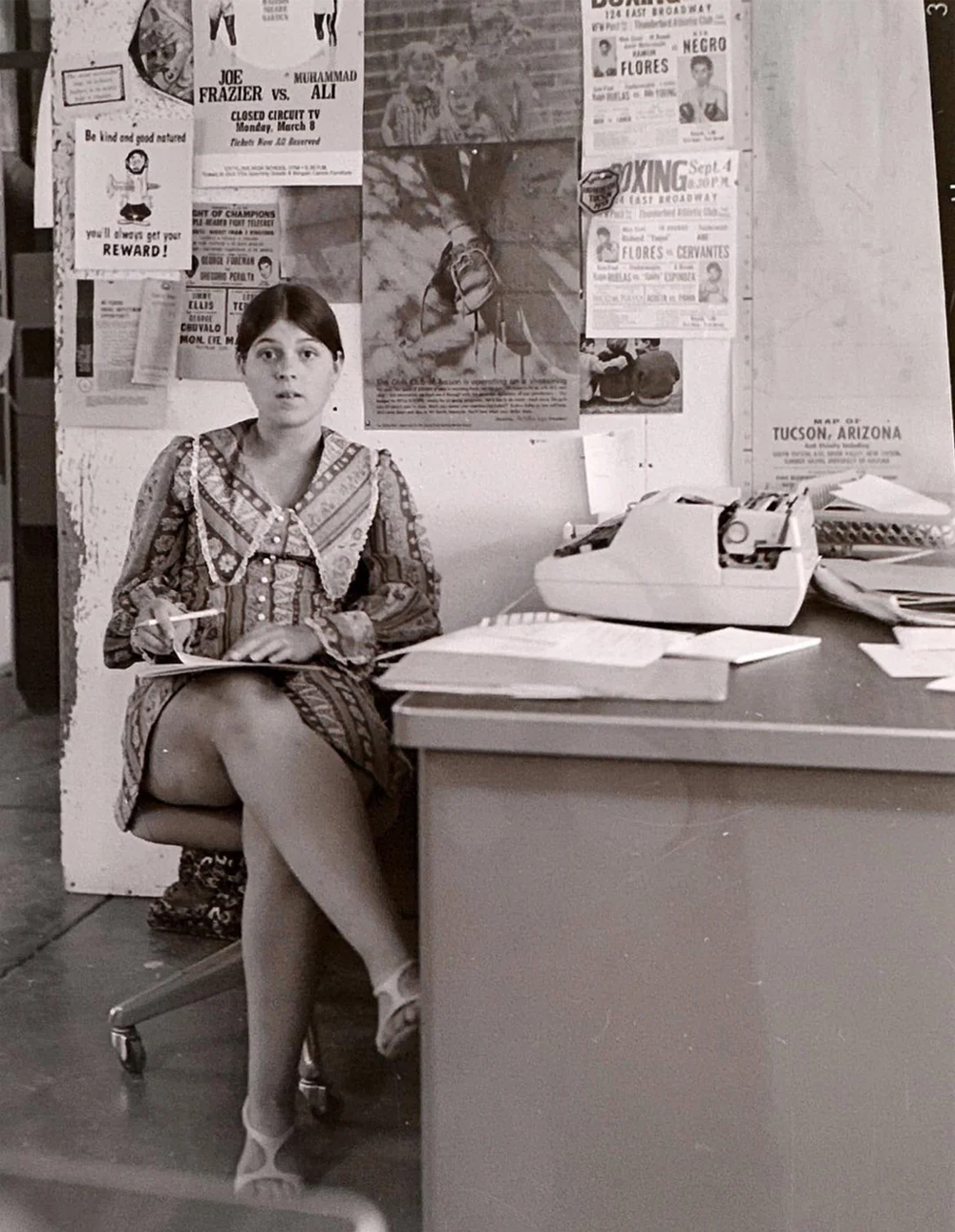 A woman in a patterned dress sits with crossed legs, holding a cup and saucer beside a desk with a typewriter. Posters and papers cover the wall behind her.