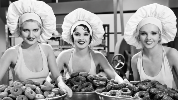 Three women in chef hats and aprons smile while holding large trays filled with donuts in what appears to be a bakery setting. The image is in black and white.