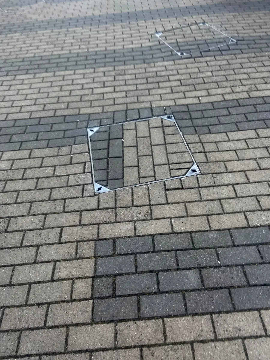 A paved sidewalk with a square maintenance cover whose bricks are misaligned with the surrounding pavement, creating a noticeable visual contrast. Another similar cover is visible further away.