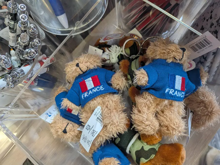 Several small teddy bears wearing blue shirts with the French flag and the word “France” are displayed in a clear plastic bin, along with some pens and other souvenirs.