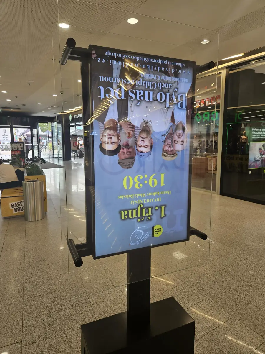 An advertisement poster displayed upside down on a digital stand inside a shopping mall, showing faces and text that are inverted.