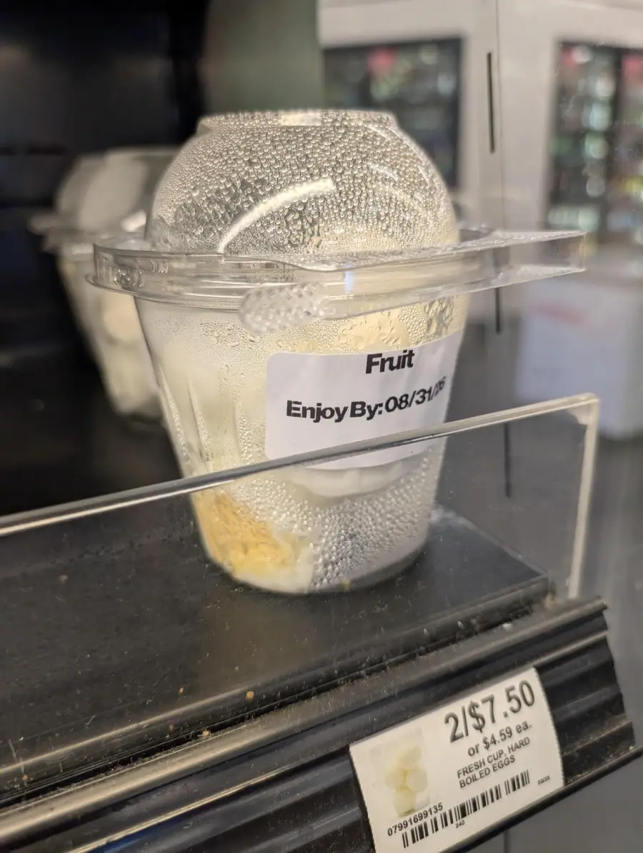 A plastic cup labeled “Fruit” with an expiration date sits on a store shelf, mostly empty except for a small piece of fruit at the bottom. Condensation covers the inside of the lid.