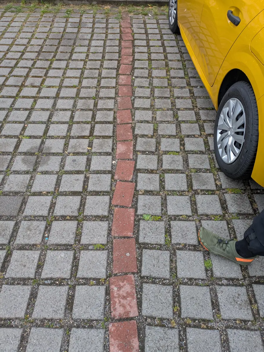 A yellow car is parked on a paved lot with a wavering line of red bricks. A person wearing green shoes and dark pants stands nearby.
