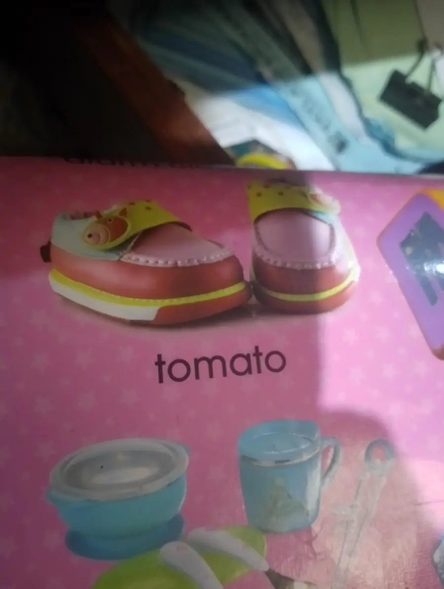 A pair of pink and yellow children’s shoes with cartoon faces on them, labeled “tomato,” shown on a pink background alongside toy kitchen items.