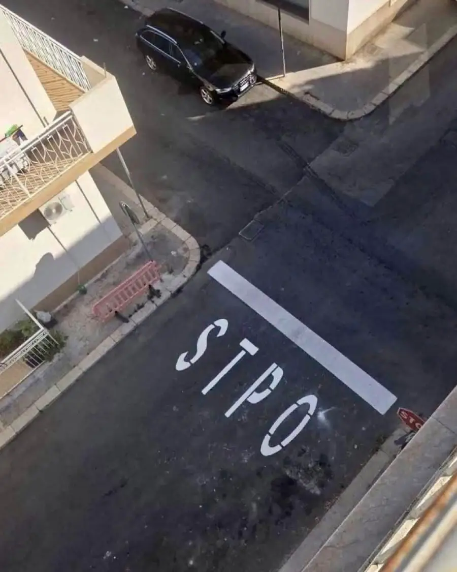 A street viewed from above shows a misspelled “STPO” painted at a stop line, with a black car parked nearby and some buildings and barriers visible.