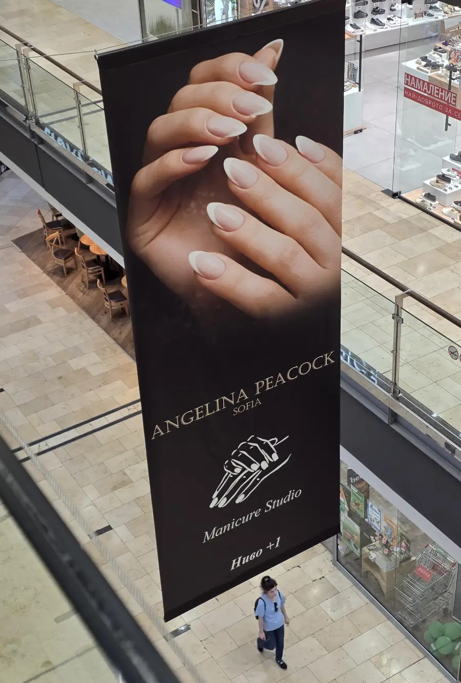 Large vertical banner for “Angelina Peacock Sofia Manicure Studio” showing manicured hands, displayed in a shopping mall. A person walks nearby on the tiled floor below.