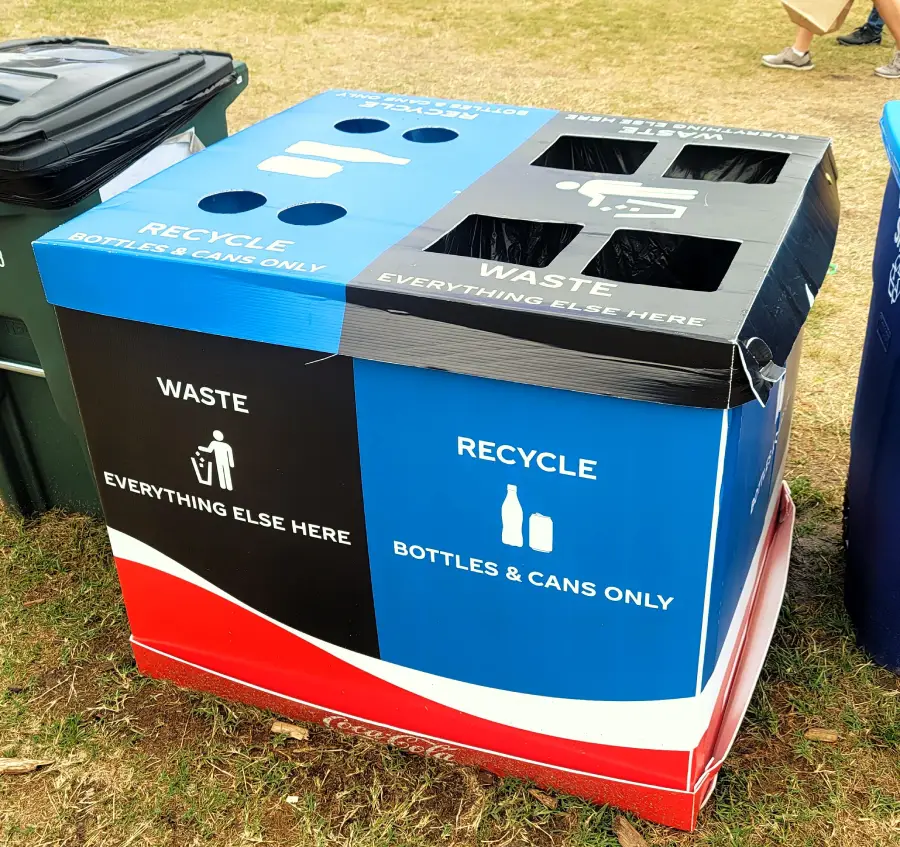 Blue and black recycling and waste bin station with labeled holes for bottles, cans, and general waste, placed outdoors on grass.