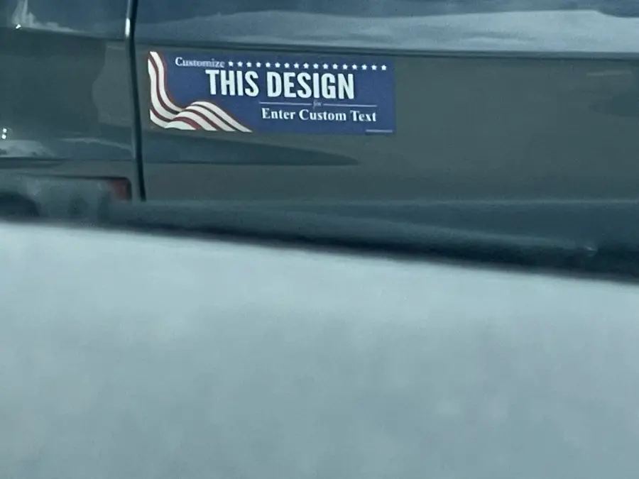 A car with a bumper sticker that says “Customize THIS DESIGN Enter Custom Text” with a stylized American flag design. The sticker appears unfinished, as if a custom message was never added.