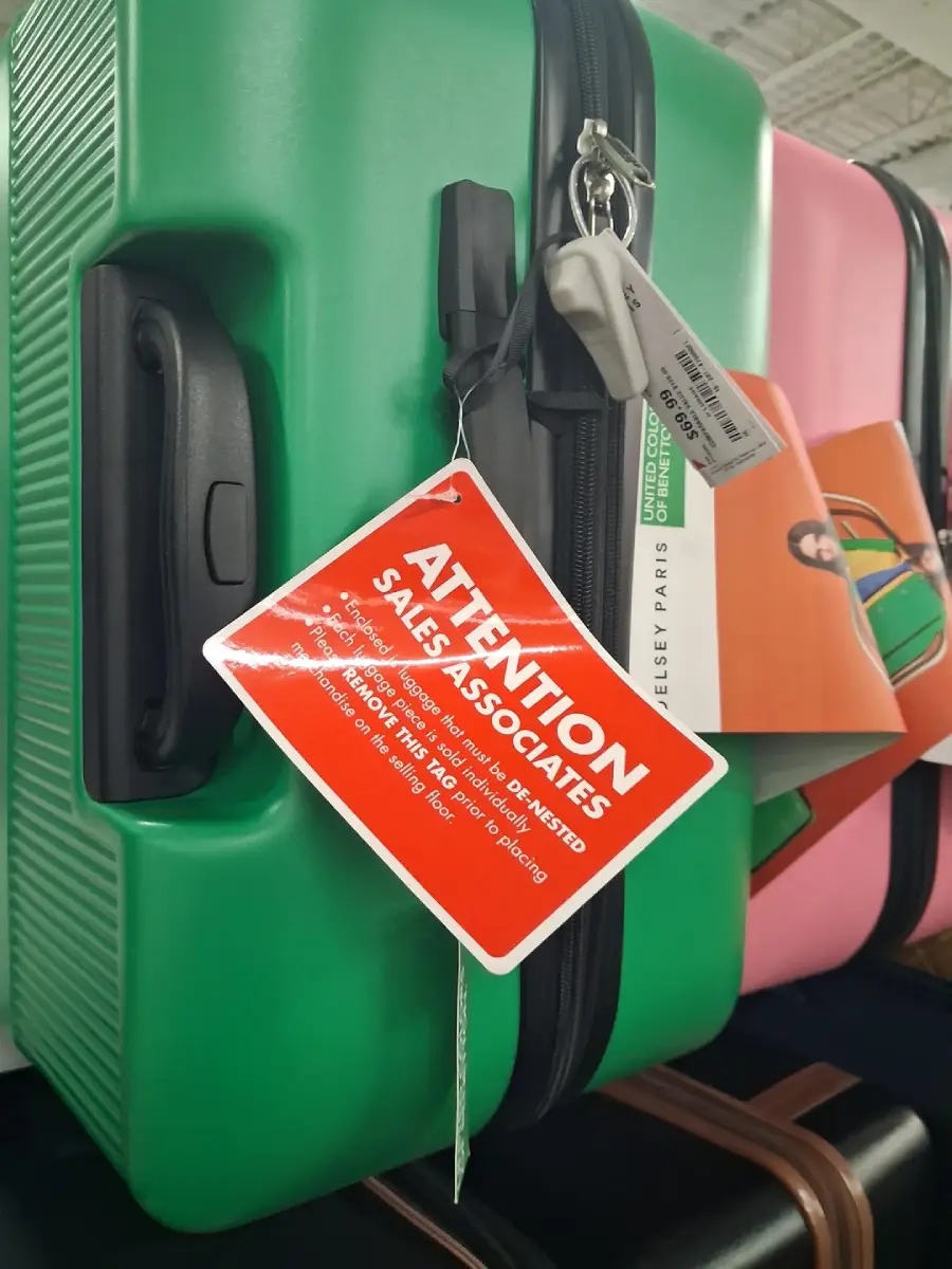 A green suitcase with a red tag reading “ATTENTION SALES ASSOCIATES” hangs on a store display rack next to another pink suitcase. The tag provides instructions to workers.