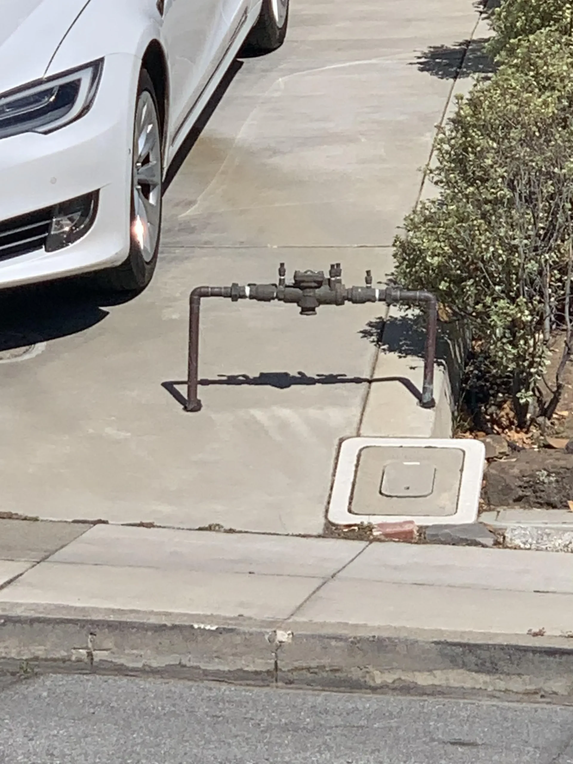 A white car is parked in a driveway next to a metal water meter assembly and shrubs; part of the meter setup extends onto the concrete near the curb.