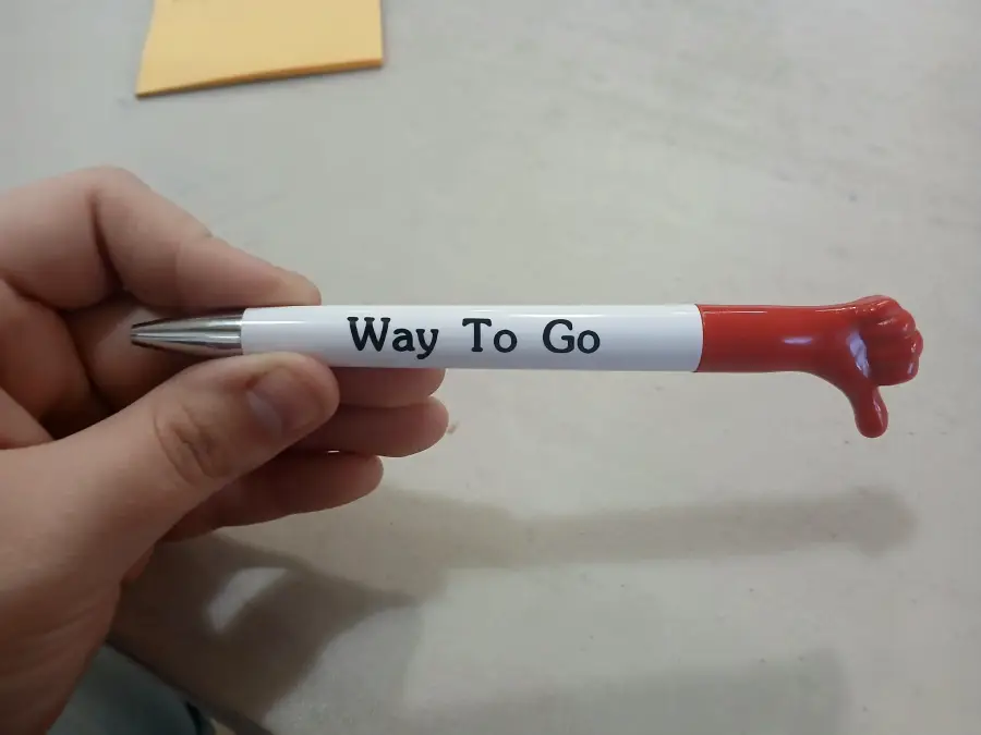 A hand holds a pen labeled “Way To Go.” The pen’s end is shaped like a red thumbs down instead of a thumb up.