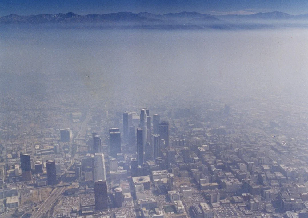 Aerial view of a city with tall buildings in the foreground, partially obscured by a thick layer of smog. Mountains are visible in the distant background under a hazy sky.