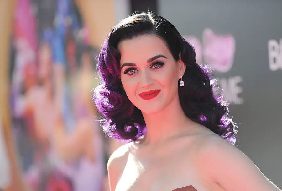 A woman with wavy dark purple hair, red lipstick, and drop earrings poses at an outdoor event, smiling at the camera with a blurred colorful backdrop behind her.