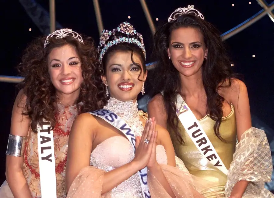Three beauty pageant winners wearing sashes and tiaras smile on stage. The center winner holds her hands in a traditional greeting gesture; the others stand close, all dressed in elegant gowns against a dark, starry backdrop.