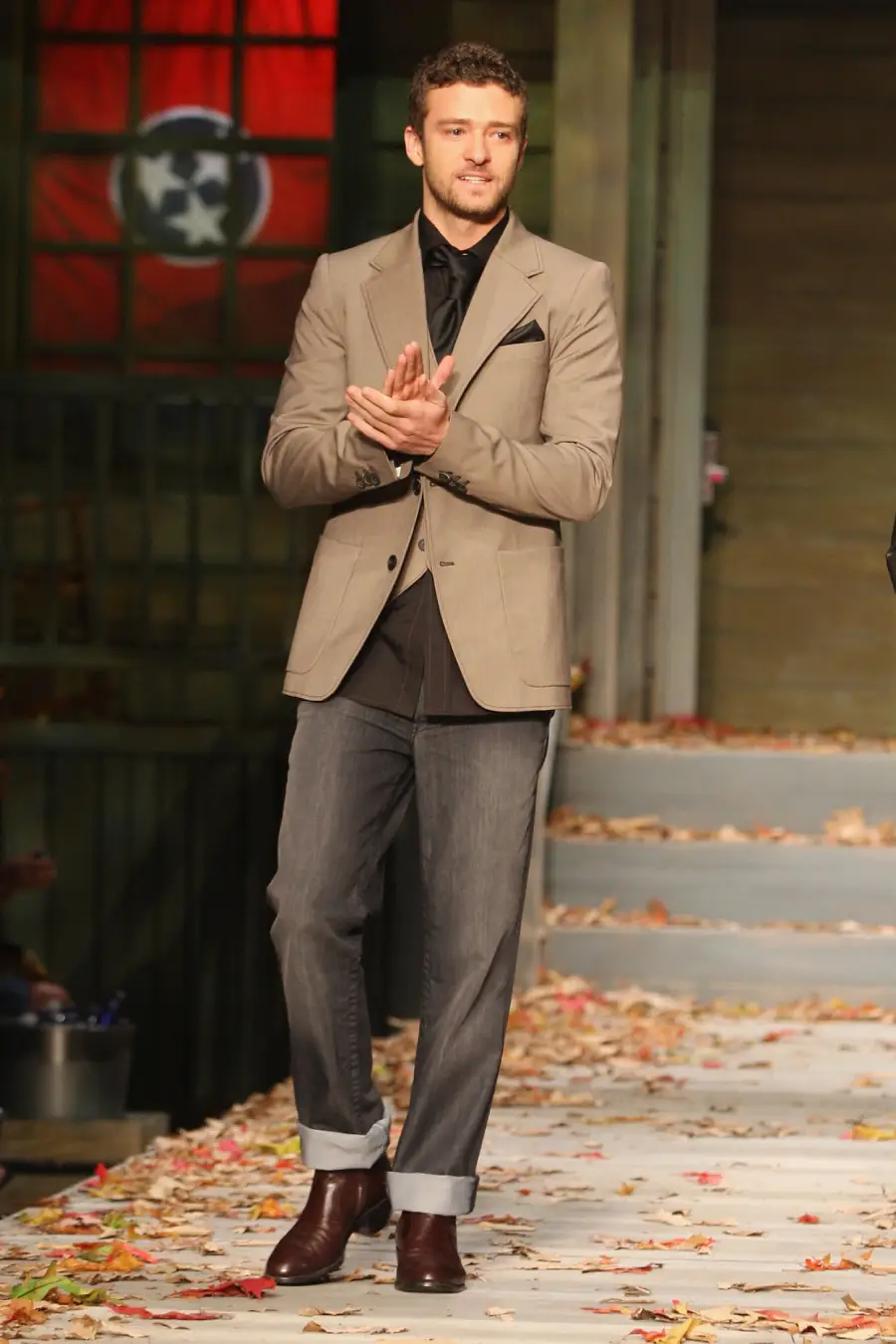 A man in a tan blazer, dark shirt, and cuffed jeans stands clapping on a leaf-strewn stage with stairs, in front of a flag featuring three white stars in a blue circle.