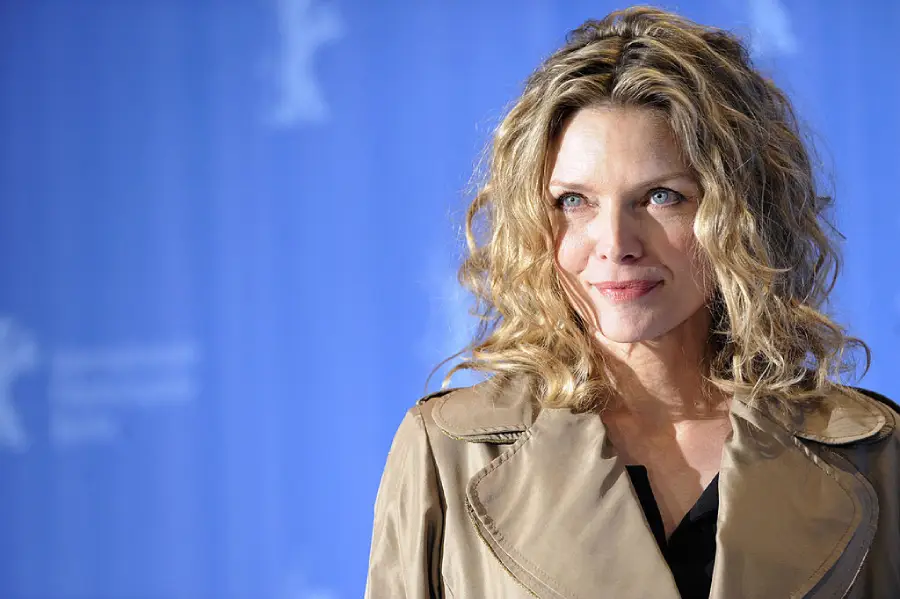 A woman with wavy blonde hair wearing a tan trench coat stands in front of a blue background, looking slightly to the side and smiling softly.