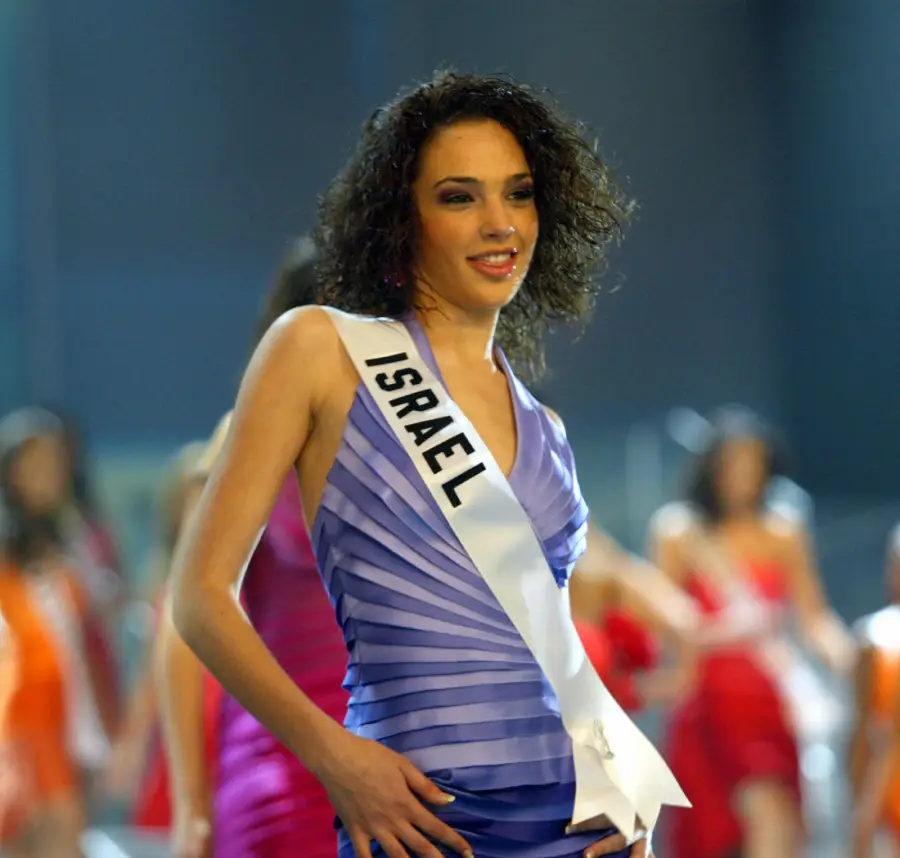 A woman wearing a sash labeled “Israel” and a sleeveless purple dress stands confidently on stage, with other contestants in colorful dresses blurred in the background.