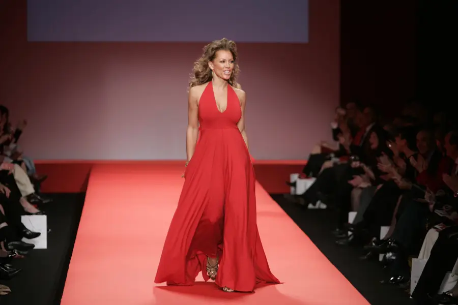 A woman walks a red runway in a flowing red halter gown and gold heels, smiling at an audience seated on both sides of the catwalk.