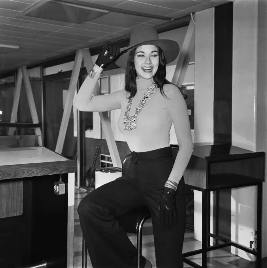 A woman in a wide-brimmed hat, turtleneck, high-waisted pants, gloves, and statement necklace poses smiling indoors, resting one hand on a chair and tipping her hat with the other.