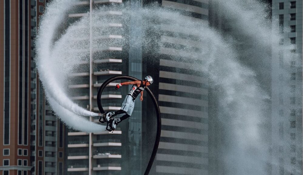 A person wearing a helmet uses a water-powered jetpack, creating a spiral spray against tall city buildings in the background.
