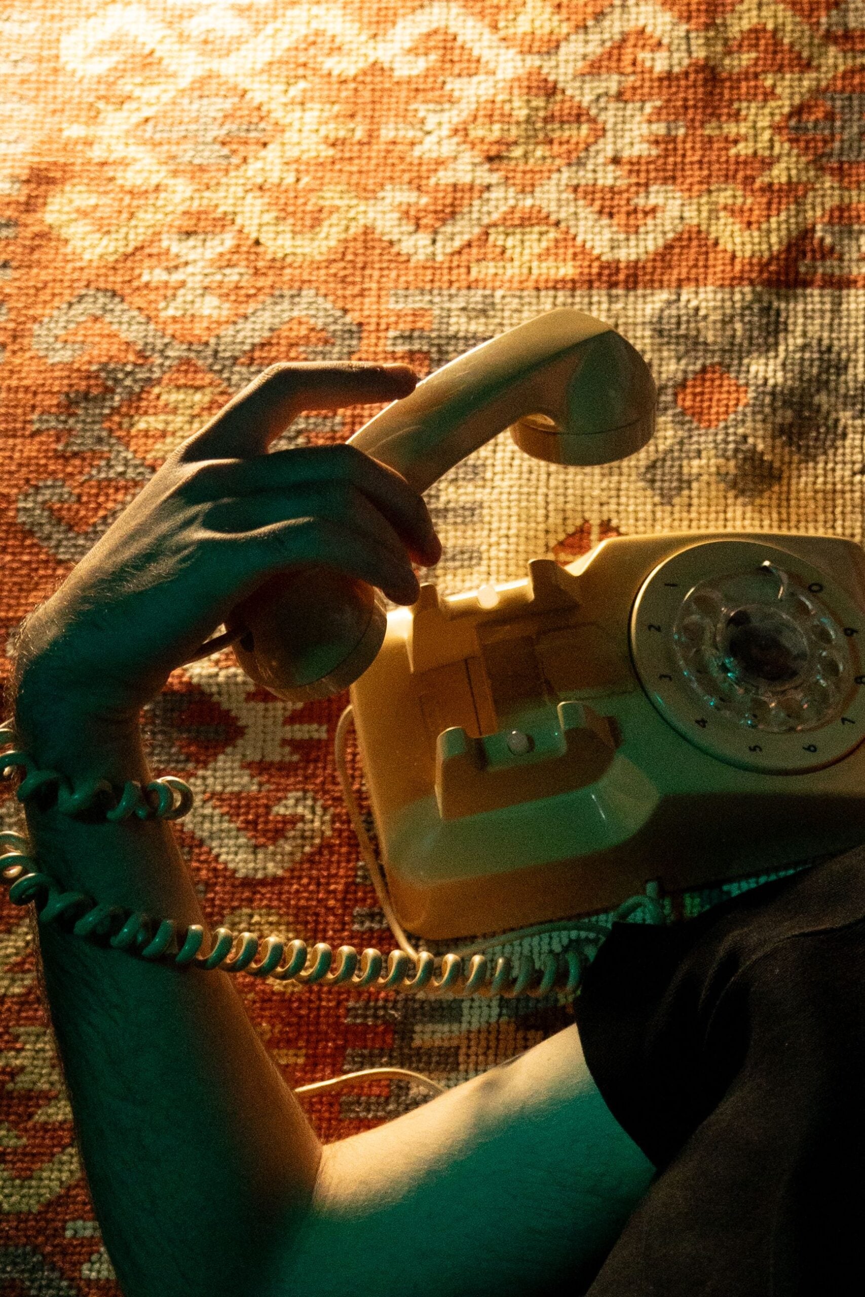 A person lies on a patterned carpet, holding a vintage beige rotary phone to their ear, with only their arm and part of their upper body visible.