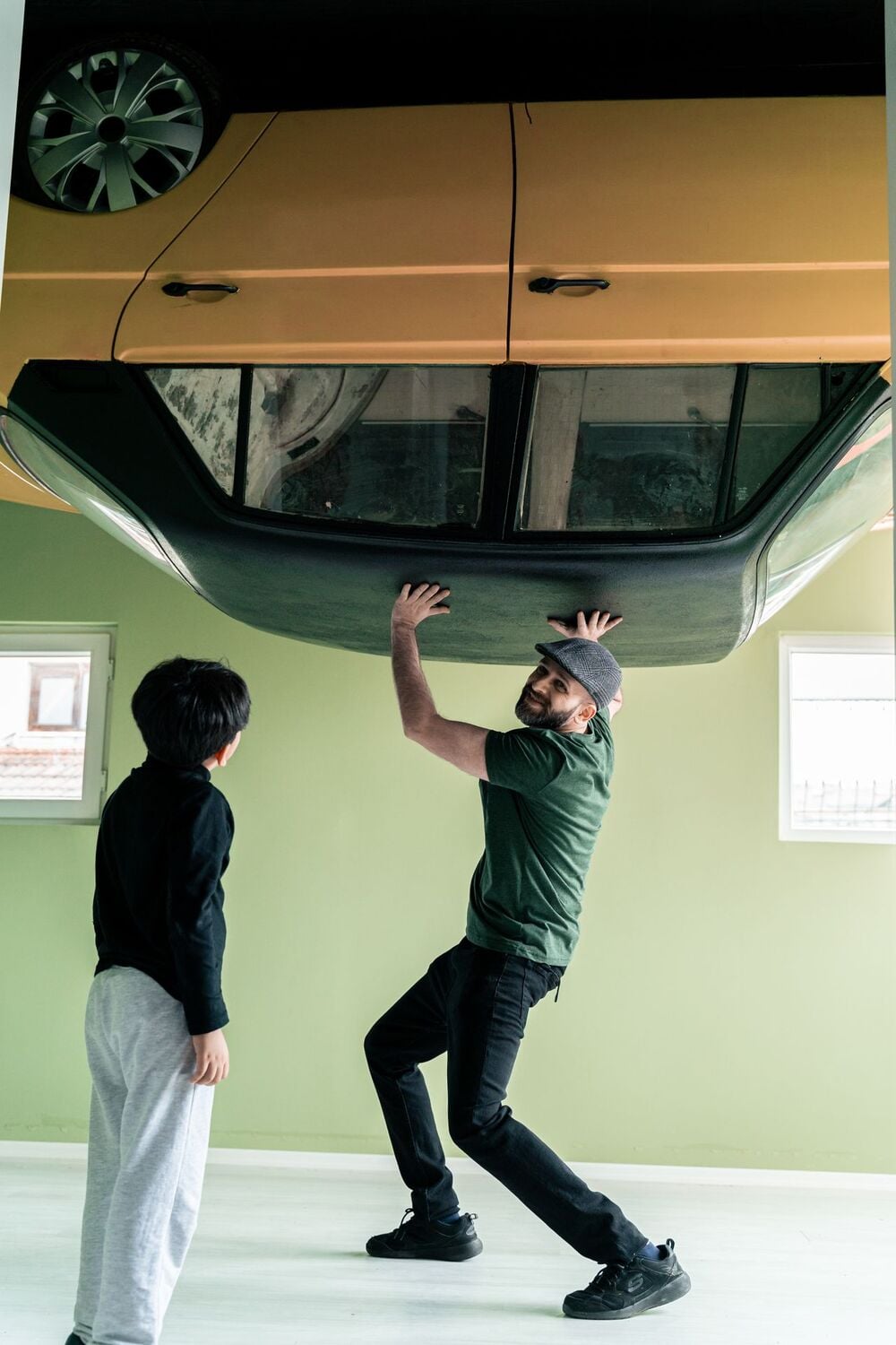A man pretends to lift an upside-down car attached to the ceiling while a child looks on in a room with green walls and two windows.