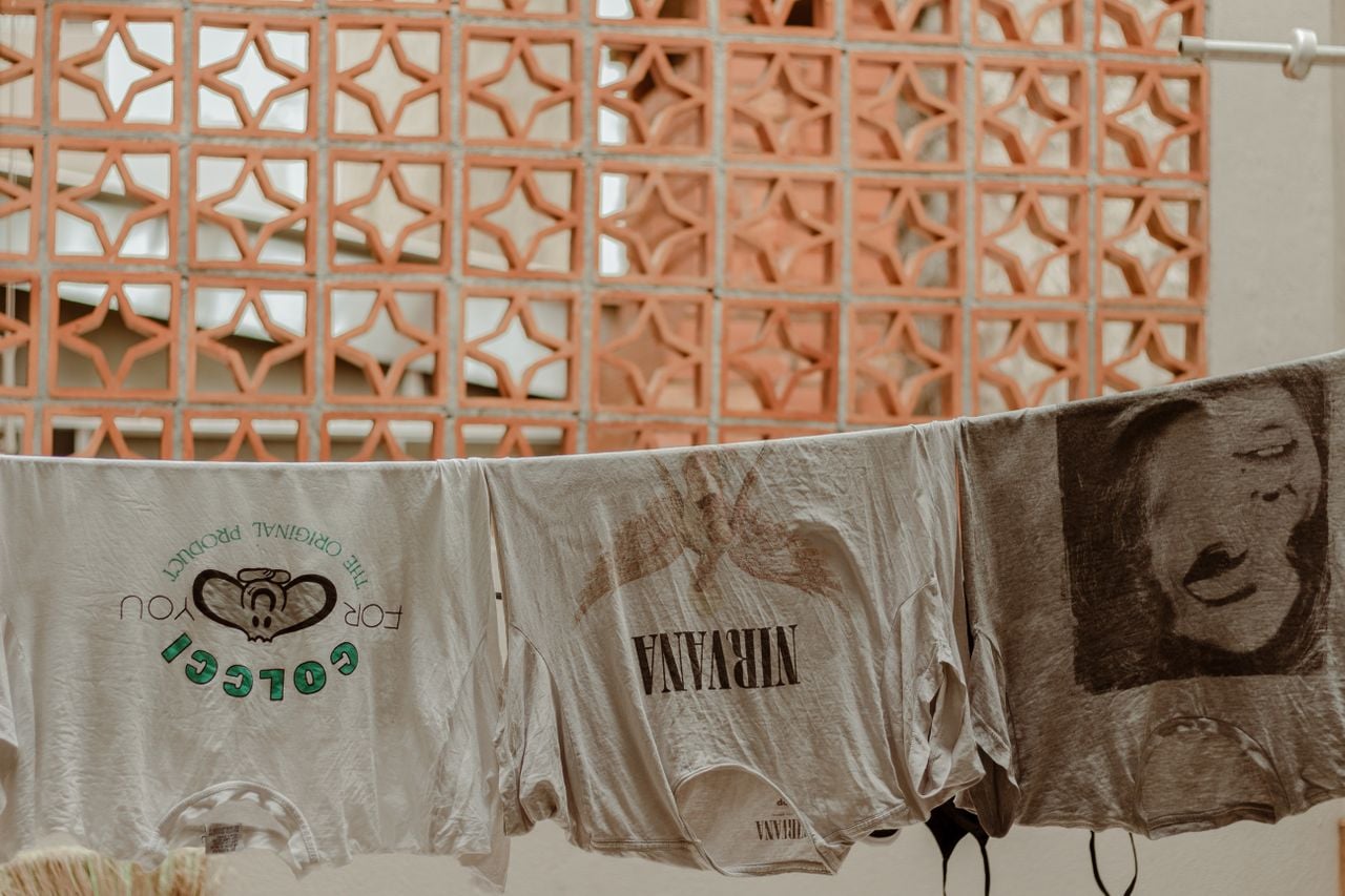 Three graphic T-shirts hang on a clothesline in front of a peach-colored brick wall with geometric cutouts. The shirts feature distinct designs and text, including “NIRVANA.”