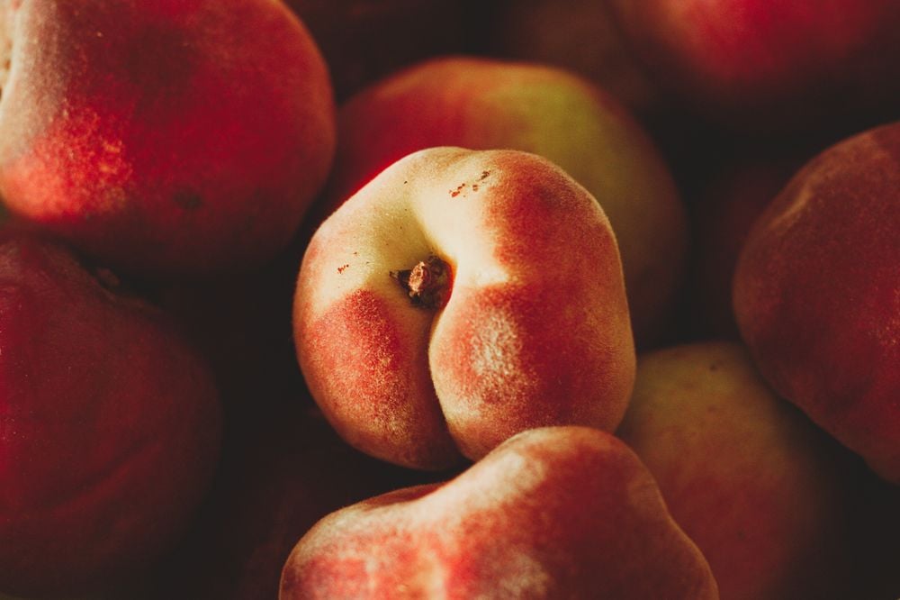 Close-up of several ripe, fuzzy peaches with red and yellow skin, focusing on a peach in the center. The lighting highlights the texture and natural imperfections of the fruit.