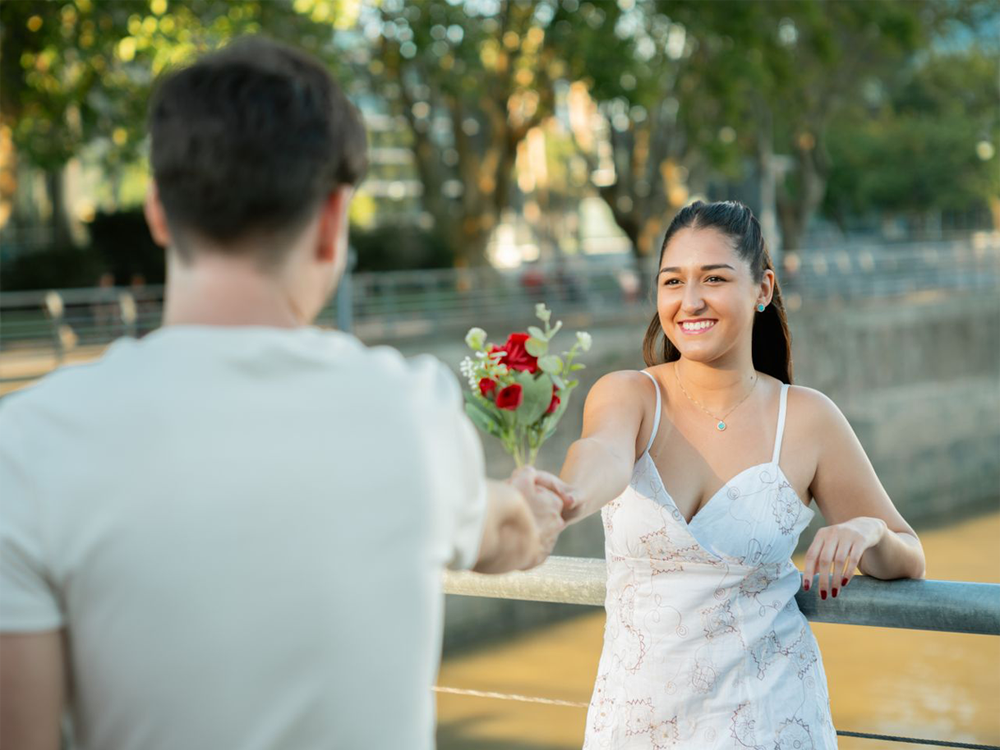 A man hands a small bouquet of flowers to a smiling woman in a white dress, standing by a riverside railing with trees and water in the background.