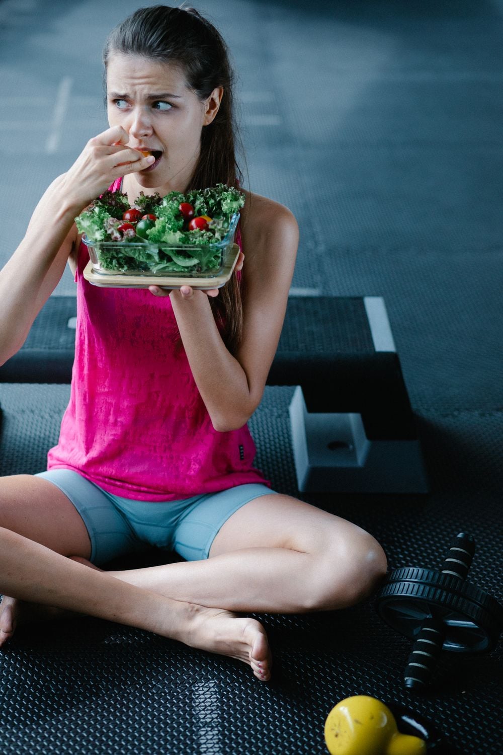 A woman in workout clothes sits on the floor, holding a salad and eating. Exercise equipment, including weights, is nearby.