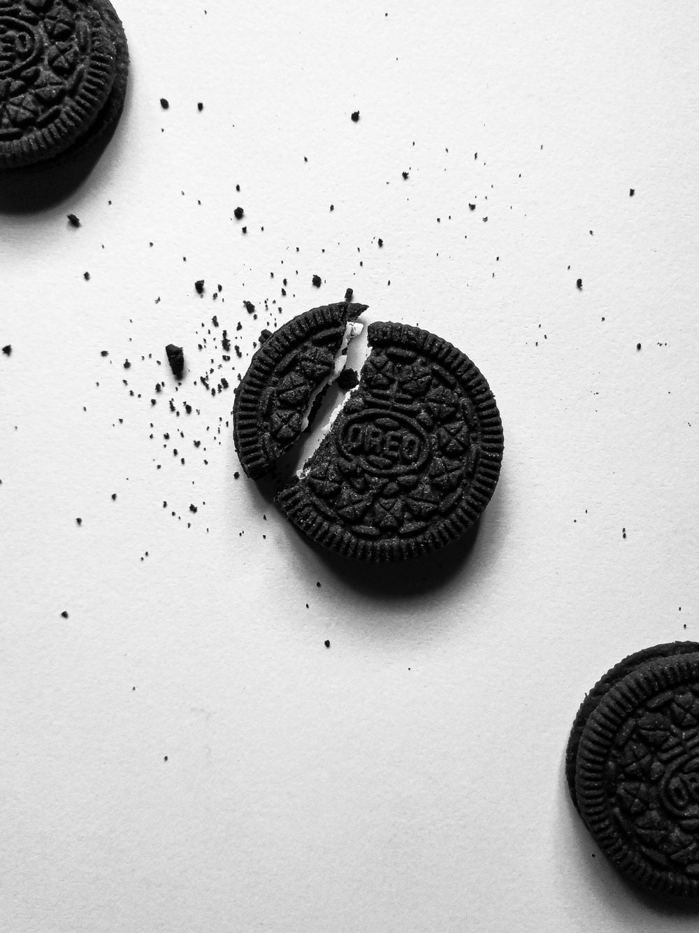 A broken chocolate sandwich cookie with cream filling is surrounded by crumbs on a white surface, with three more cookies partially visible at the edges.
