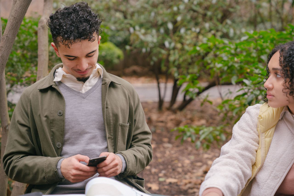 A young man looks down at his phone while sitting on a bench outdoors, and a young woman beside him looks at him. The background is filled with green foliage and trees.