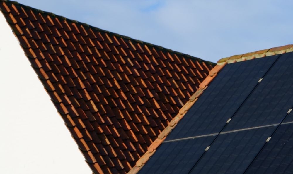 Close-up of two rooftops: one with traditional red tiles, the other with solar panels installed, against a background of blue sky with some clouds.