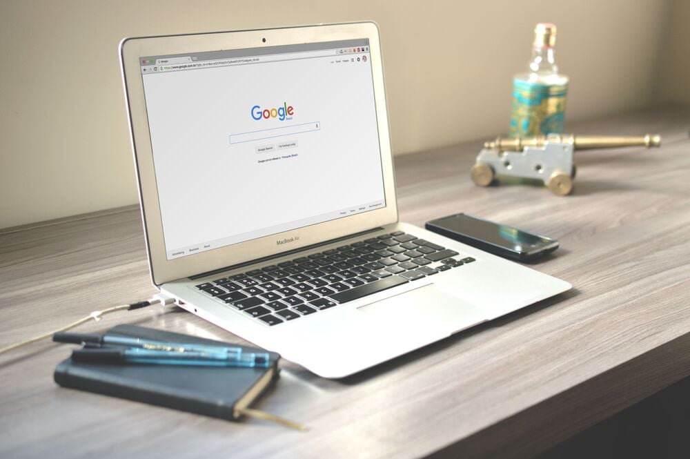 A MacBook on a desk displays the Google homepage. Nearby are a smartphone, a closed notebook with pens, earphones, a decorative cannon, and a glass bottle. The setting appears neat and organized.
