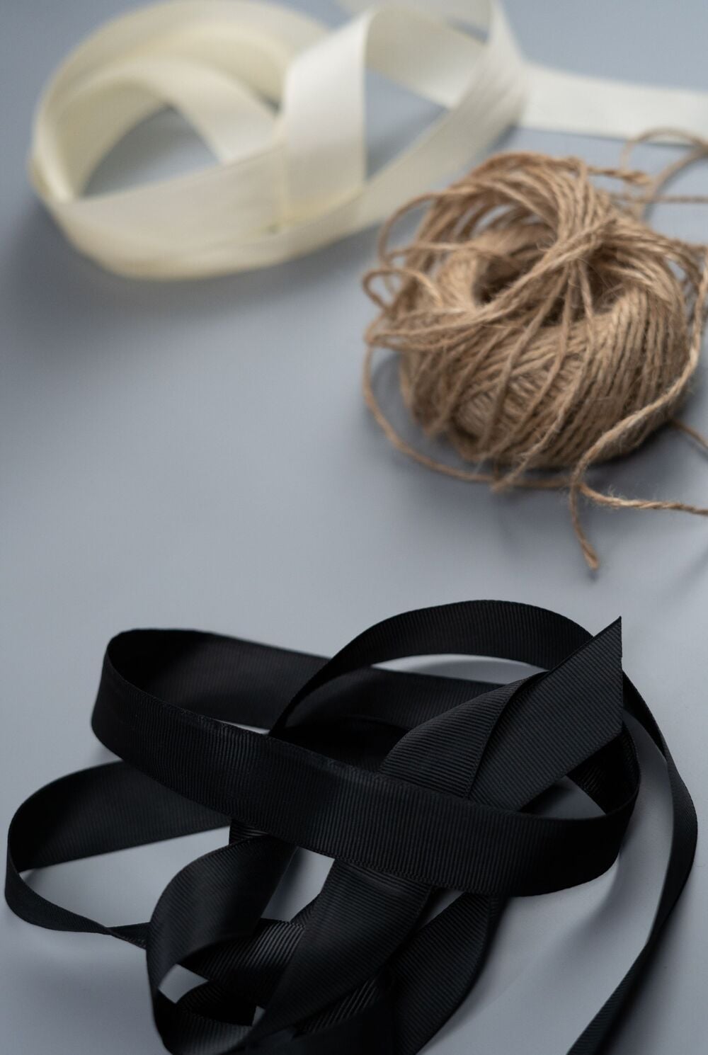 A spool of brown twine, a looped cream ribbon, and a looped black ribbon are arranged on a gray surface.
