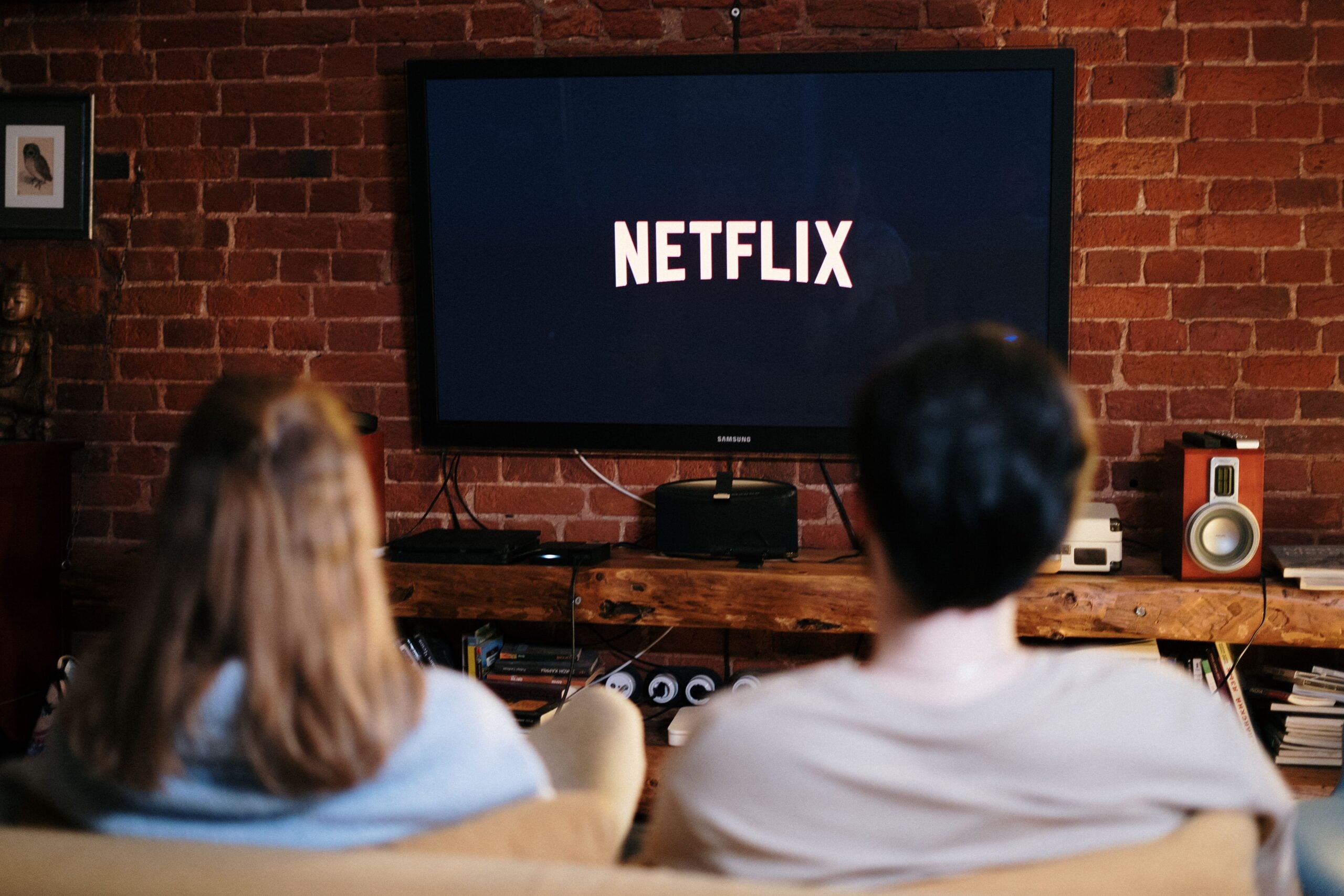 Two people sit on a couch facing a TV displaying the Netflix logo in a cozy living room with a brick wall, wooden shelf, and speakers.