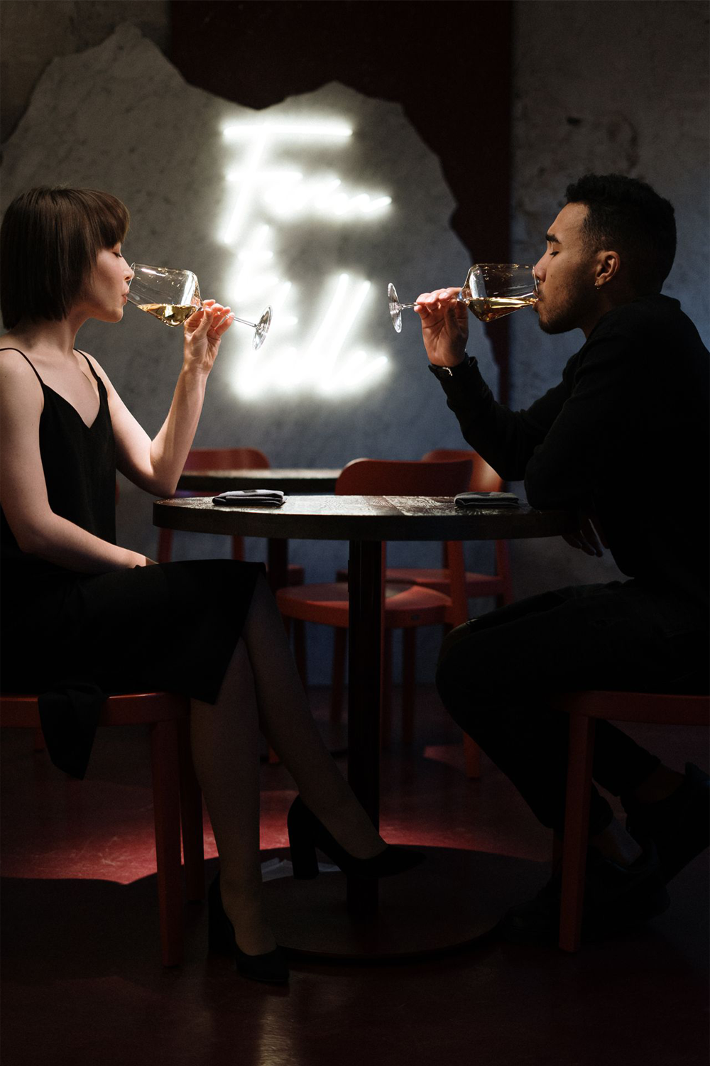 A woman and man sit across from each other at a small table, dressed formally, sipping wine. The background features illuminated, blurry white text on a rough wall.