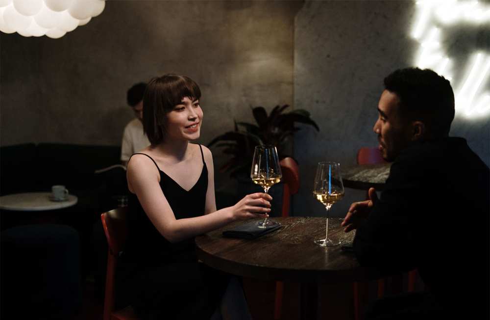 Two people sit at a dimly lit restaurant table, smiling and talking while holding glasses of white wine. A soft light hangs overhead, and another patron is seated in the background.