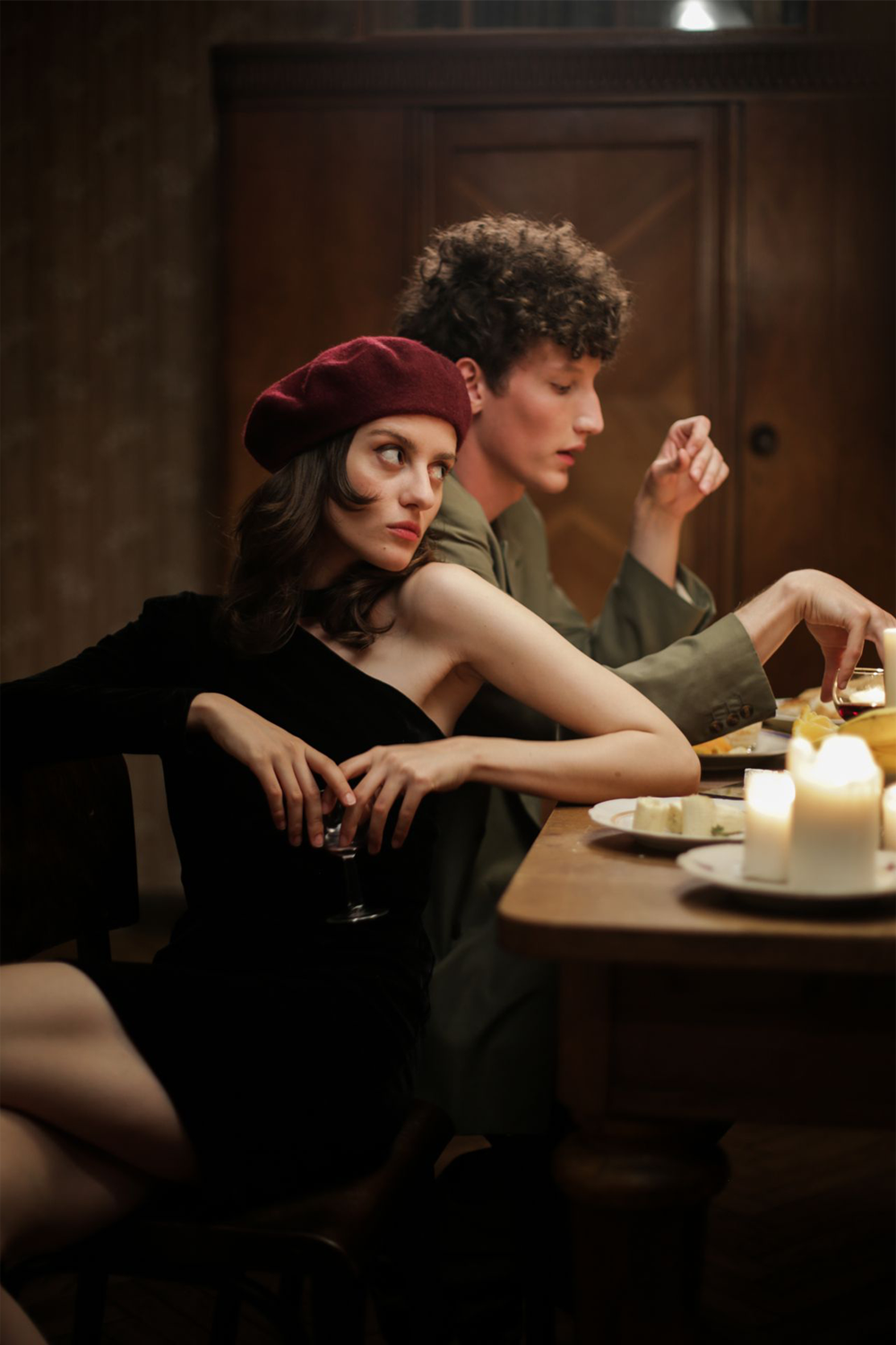 A woman in a black dress and red beret sits at a dinner table with a man, both holding drinks. Lit candles and food are on the table. The woman looks away, appearing thoughtful or distant.