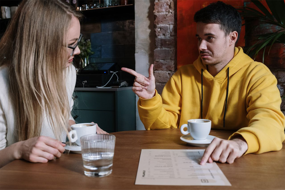 A woman and a man sit at a table with coffee cups. The man, wearing a yellow hoodie, points at a menu while talking to the woman, who looks at him attentively. A glass of water and a menu are on the table.