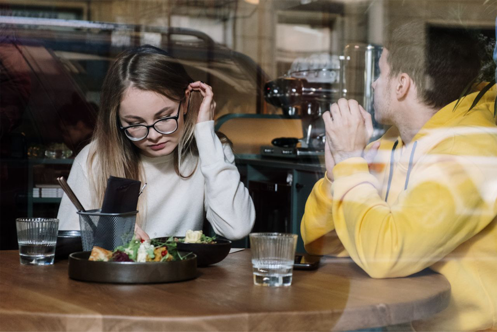 A woman with glasses looks down at her food while a man in a yellow hoodie sits across from her, both at a wooden table with drinks and salad in a cafe, seen through a window.