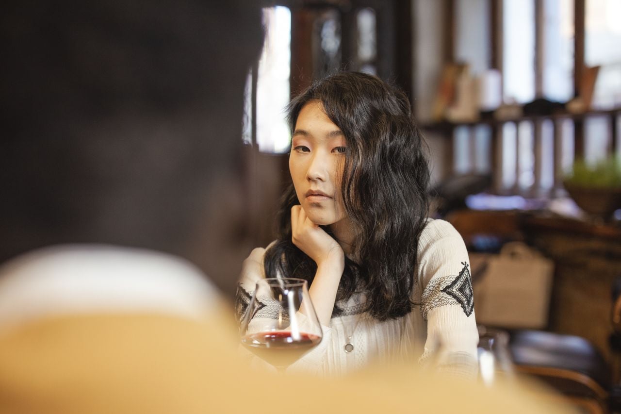 A woman with long dark hair sits at a table, resting her chin on her hand, looking thoughtful. A glass of red wine is in front of her, and another person is blurred in the foreground.