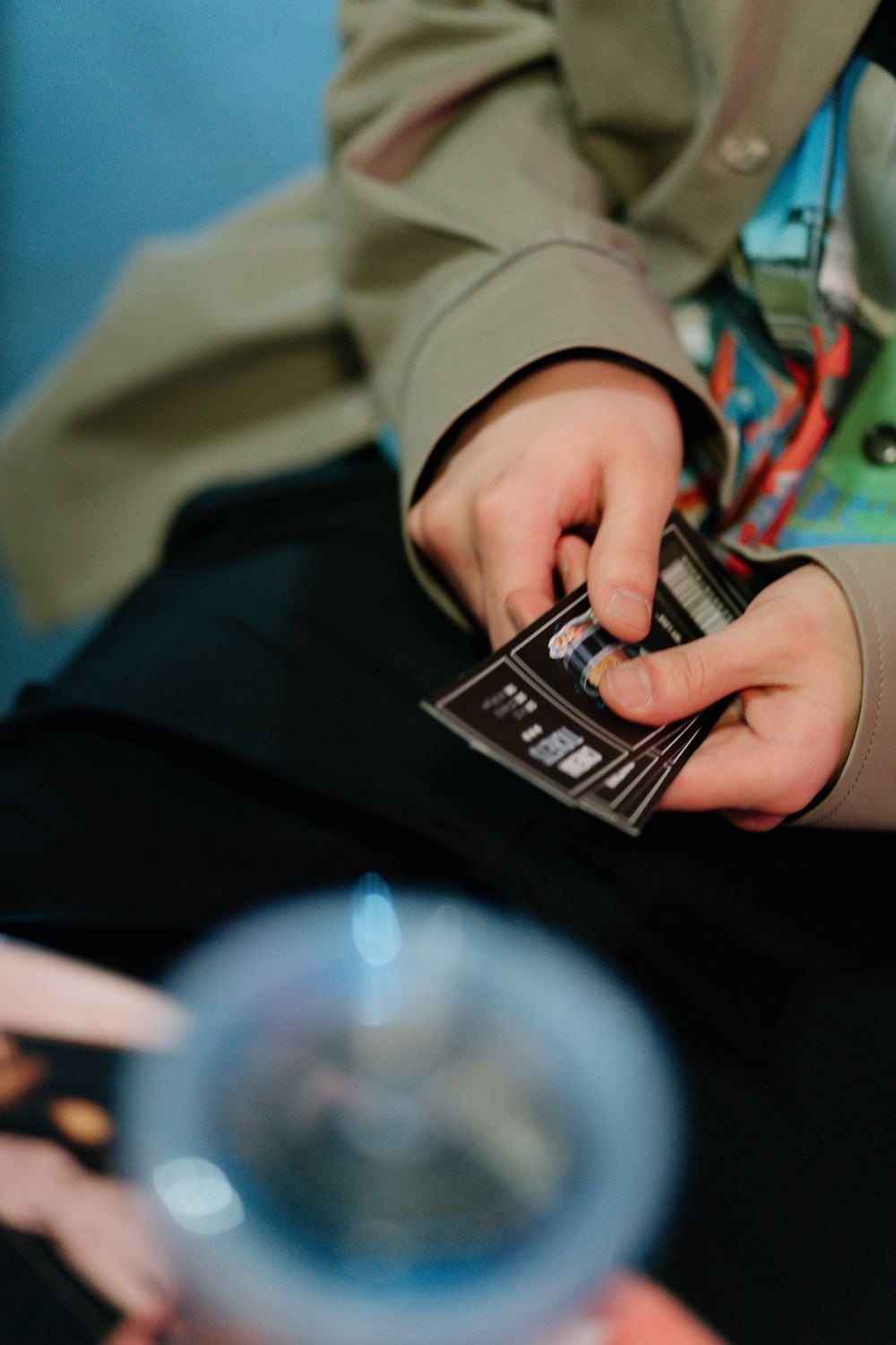 A person wearing a beige jacket is holding and sorting black trading cards. A blurred drink cup is visible in the foreground.