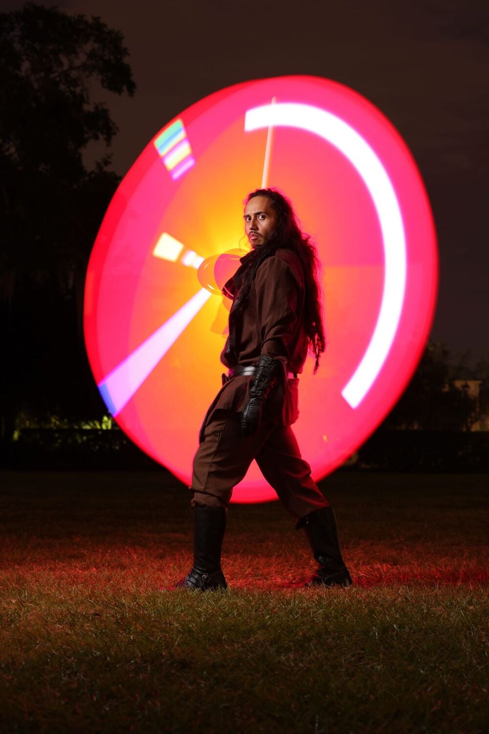 A person in brown costume poses outdoors at night, holding a large red circular LED light that glows brightly behind them, creating a vivid sci-fi effect on the grass and surroundings.