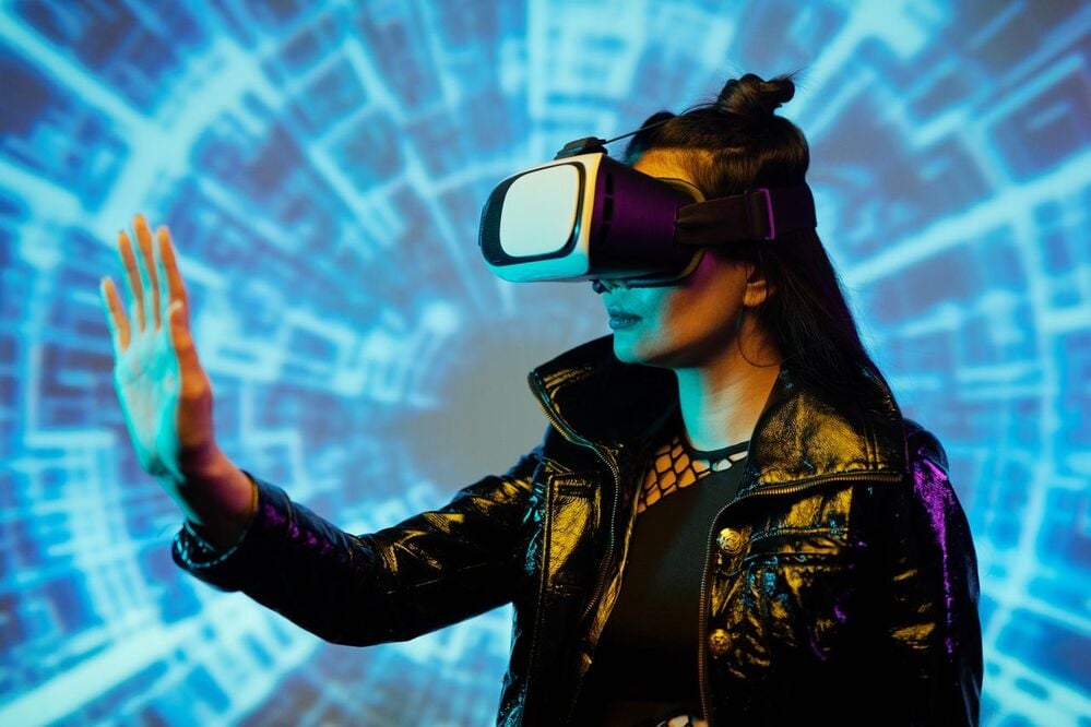 A person wearing a VR headset and black jacket reaches out with one hand, with a blue digital, tunnel-like background suggesting virtual reality or futuristic technology.