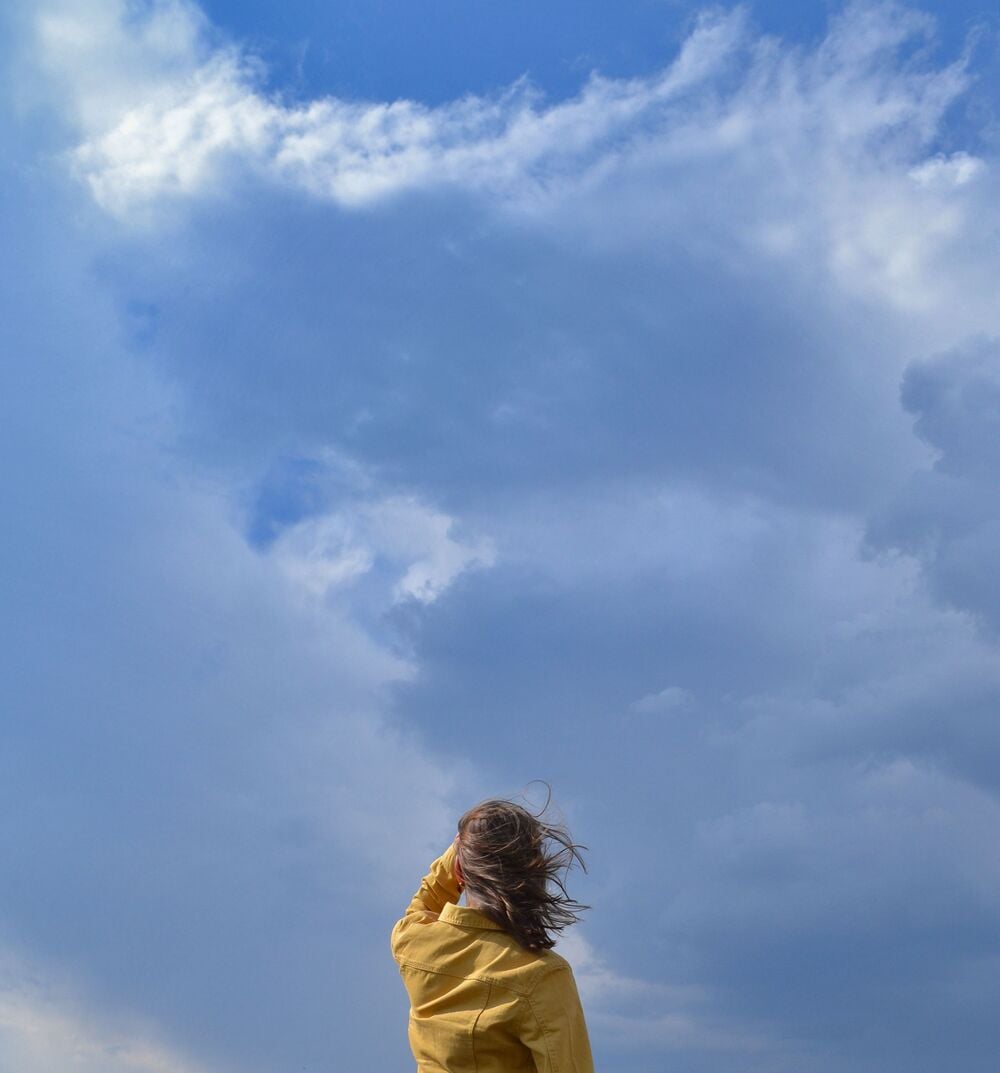 Person in a yellow jacket stands with back to camera, looking up at a cloudy blue sky. Their hair is blowing in the wind, and the sky is filled with large, white clouds.