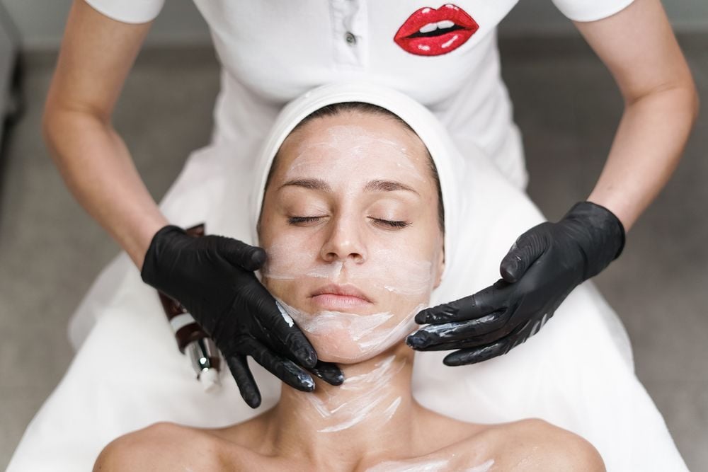 A woman with a headband is lying down with her eyes closed while a person in black gloves applies a facial mask to her face. The person wears a white shirt with a red lips design on the chest.