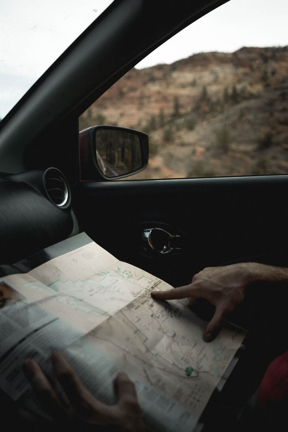 A person sits in a car holding and pointing at a paper map, with a scenic, rocky landscape visible through the window.